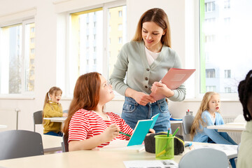 Smiling teacher talking to schoolgirl while standing near desk in the classroom