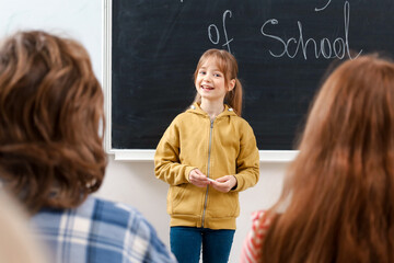 Smiling girl holding chalk standing near the blackboard and talking to classmates. Back to school