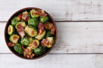 Delicious roasted Brussels sprouts and bacon in bowl on light wooden table, top view. Space for text