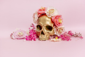 A golden skull surrounded by dried flowers on a soft pink surface
