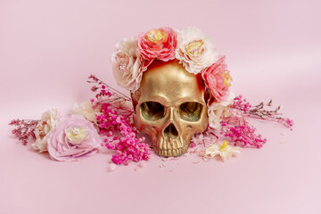 Front image of a golden skull surrounded by dried flowers on a soft pink surface