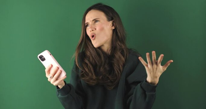 Mad and shock young brunette beautiful woman using mobile phone isolated on green background. Yelling unhappy woman hold mobile phone. Disappointed sad upset lady horrified impressed news. WTF. Oh no