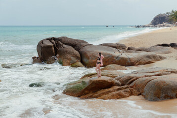 Woman strolls along a boulder-strewn coast with waves gently crashing in and turquoise sea extending to the horizon