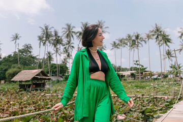 Smiling woman stands on a wooden walkway by a lotus pond, with palm trees in the background