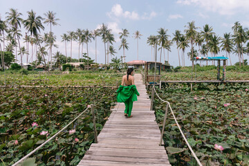 Woman strolls on a bridge over a lotus-covered pond, surrounded by palm trees under a cloudy sky