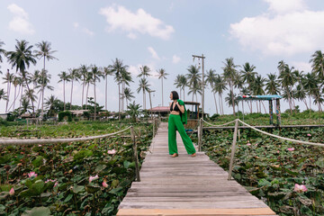 Woman strolls on a rustic boardwalk surrounded by lush greenery and lotus ponds, with tall palm trees standing against the sky
