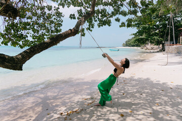 Woman enjoys a peaceful swing on a rope tied to a tree, against the backdrop of a tranquil beach and clear skies
