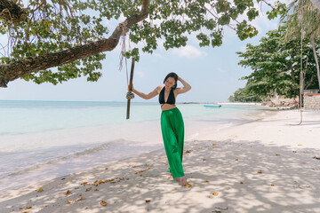 Woman in a black top and green pants stands by a tree on a sunny beach with a white sand backdrop