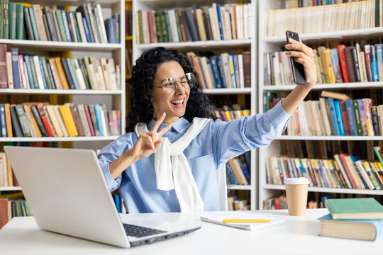 Cheerful Hispanic female student takes a playful selfie among books in a library setting, exemplifying student life and happiness.
