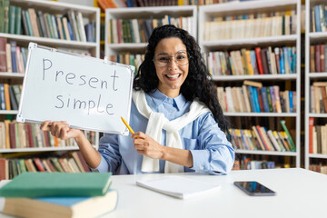Cheerful educator teaching the present simple tense, using a whiteboard in front of a bookshelf-filled library.