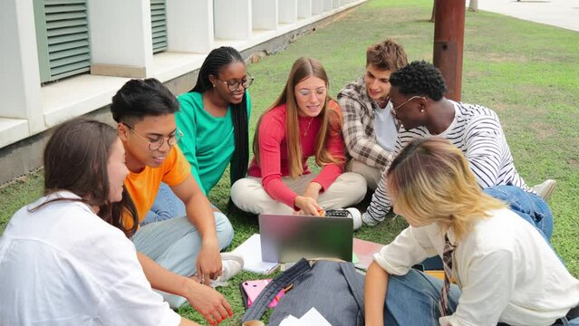 Multiracial students using a laptop computert sitting in a circle on the grass, doing homework outside at university campus. Young high school people working together on a academy task or schoolwork