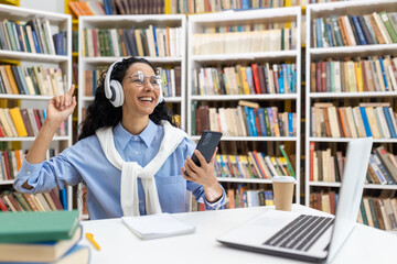 A cheerful student is listening to music on headphones, using a smartphone in a library, surrounded by books and a laptop.