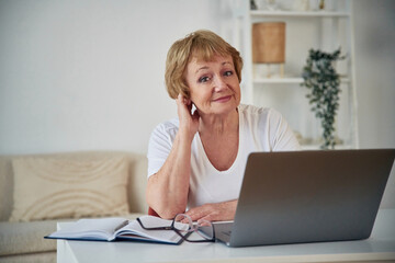 Front view. Sitting by table with laptop. Senior elderly woman is at home in the living room