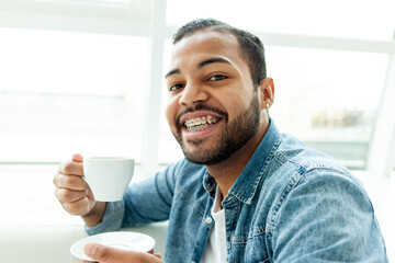 African American man with braces drinks coffee from a cup and smiles in a white cafe, a man with...