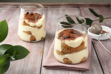 Delicious tiramisu with coffee beans in glasses and green leaves on wooden table