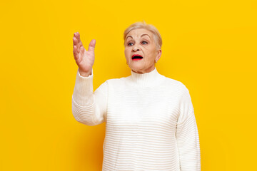 cheerful old grandmother in a white sweater announces and speaks on a yellow isolated background,...