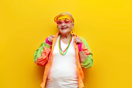 funny crazy granny in hipster clothes, glasses and cap on a yellow isolated background, elderly cool woman in fashionable youth clothes smiling