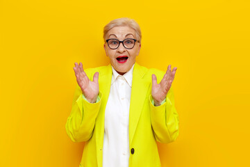 surprised old businesswoman in glasses and formal wear looks away in shock on yellow isolated background, elderly pensioner grandmother in blazer worries in amazement - obrazy, fototapety, plakaty