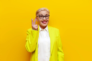 old businesswoman in glasses and formal wear showing peace gesture and tongue out on yellow...