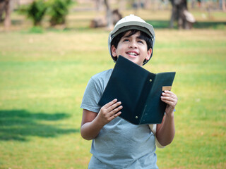 Cute Indian child wearing a white hard hat holding a sketch of a treehouse exploring a suitable area for installation..