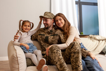 Ukrainian army soldier in camouflage uniform returned home to his family, military cadet sits on the sofa with his wife and daughter and smiles, child hugs veterans father