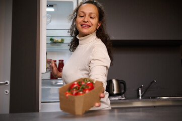 Smiling woman putting on the table cardboard box with healthy organic tomatoes cherry, standing by a fridge in the home kitchen. People. Healthy diet. Food and drink consumerism