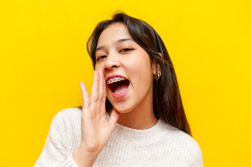 young cheerful asian woman with braces announces information and tells a secret on a yellow isolated background, korean girl speaks and whispers copy space