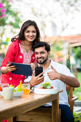 Asian Indian young couple using smartphone and tablet pc while sitting at breakfast table outdoors