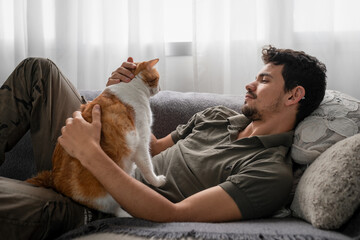 young man lying on a sofa hugs a brown and white cat
