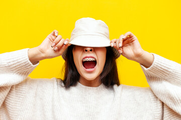 young asian woman with braces screams and covers her face with panama hat on yellow isolated...