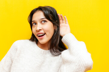 young curious asian woman with braces eavesdropping and listening on yellow isolated background,...