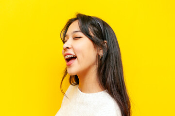 young asian woman with braces winks and smiles on yellow isolated background, korean girl hints and...