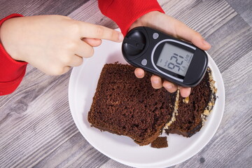 Hand of child holding glucose meter with result sugar level, sweet chocolate cake. Nutrition during diabetes