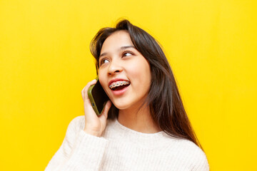 young shocked asian woman with braces talking on phone over yellow isolated background, korean girl...