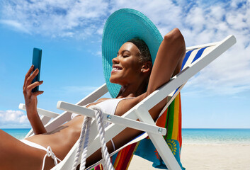Happy young woman is sunbathing on a beach deck chair, uses mobile phone, on a sunny day by the seaside, concept of a summer beach holiday, online shopping, booking travel, and resort accommodations