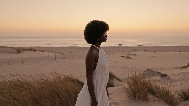 Elegant black woman in a flowing white dress stands pensively on the beach at dusk, with soft sunset light and gentle seaside tranquility.