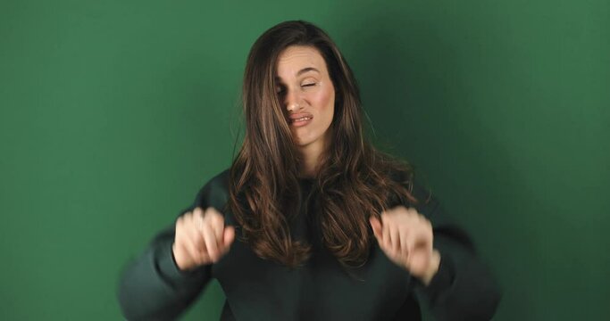 Displeased brunette young woman posing isolated on green background studio. Woman in green sweatshirt looking camera with finger gesture showing thumb down, tongue out.