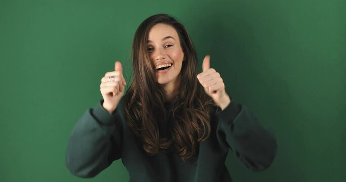 Optimistic brunette hair woman makes thumbs up gives approval shows excellent gesture says nice job gives positive feedback shares good opinion, wow, isolated over green background.