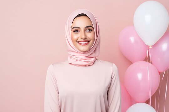 A woman wearing a pink hijab is smiling in front of a bunch of pink balloons