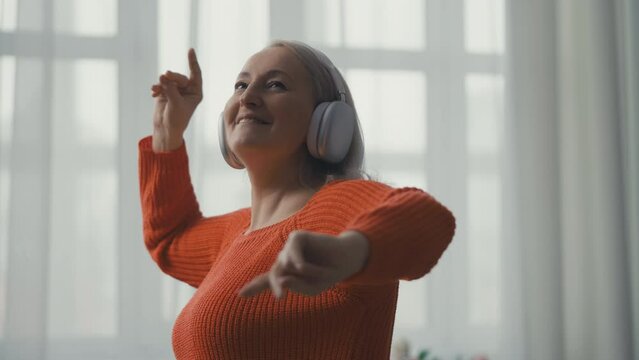 Happy middle-aged woman in headphones dancing and smiling at home, good mood