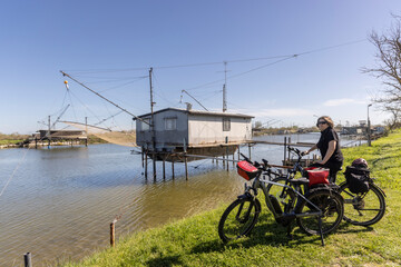 Woman cycles on an e-bike through the Po Delta in Northern Italy. In the background is a typical Italian pile dwelling, called Trabucco