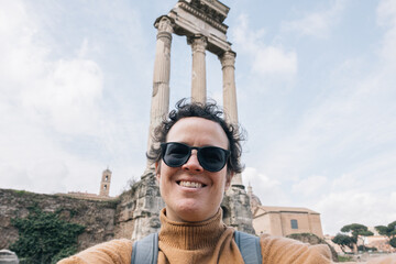 A  woman with sunglasses and backpack is taking a selfie in the Roman forum, she is smiling and...