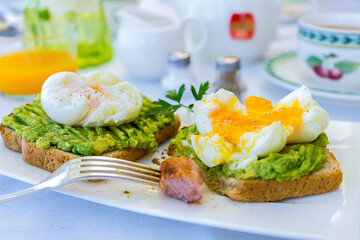 Fresh smashed avocados on wholemeal toast topped with organic poached eggs with a piece of sausage on a fork