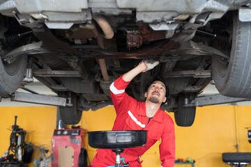 Male mechanic working at garage. Asian male mechanics checking car engine underneath lifted car at auto car repair service. Car service and Maintenance concept - obrazy, fototapety, plakaty