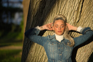 Portrait of woman, young, pretty, blonde, with eyes closed and denim dress, with her hands behind her head, leaning on the trunk of a large tree receiving the sun's rays at sunset. - Powered by Adobe