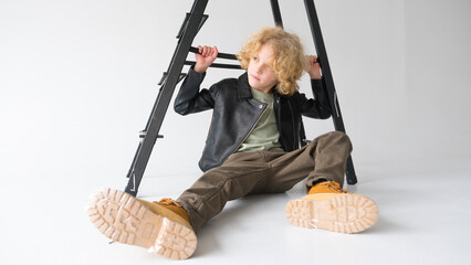 Young Boy With Curly Hair Sitting Under a Black Ladder on a White Background