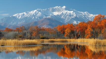   A mountain range mirrors in the tranquil water of a lake encircled by tall grass and trees with orange leaves