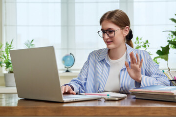 Young female college student sitting at desk, talking in web camera of laptop