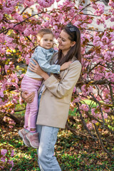 Young mother and daughter are standing in the park near a sakura tree that has blossomed with pink flowers in the spring in Prague.