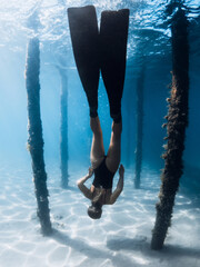 Woman freediver under the pier in blue ocean. Female swims with fins dives between the pier pillars